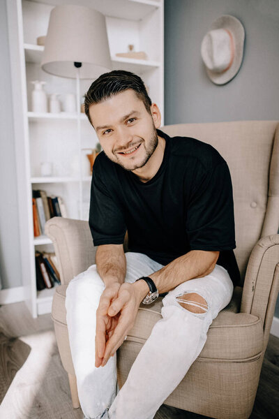 Portrait of an attractive smiling young bearded man wearing casual clothes sitting on a couch at the living room