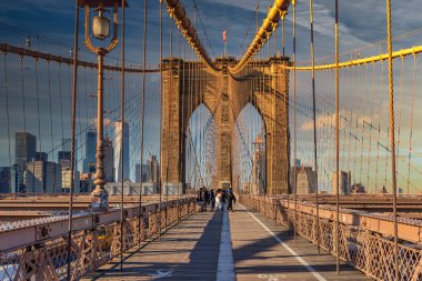 Brooklyn Köprüsü gündüz manzarası, insanlar yürüyor, Amerikan bayrağı en tepede ve ufuk çizgileri arka planda.