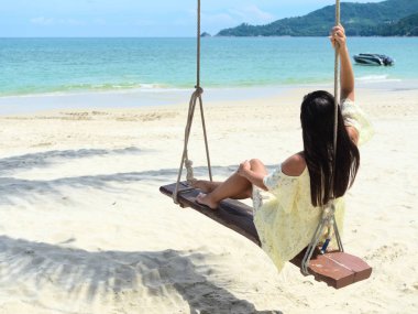 Güneşli bir günde salıncakta tek başına oturan ve denize doğru bakan bir kadın.