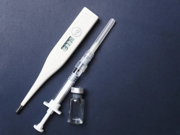 Thermometer showing a temperature of 37.7. A syringe with a needle filled with medicine, an empty vial of the drug. Top view