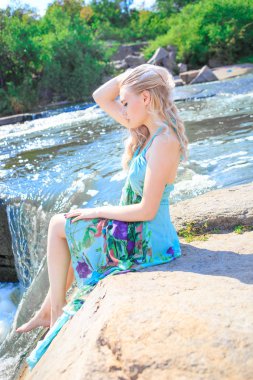 Genç sarışın kaynayan bir nehrin kıyısında elbise giymiş.