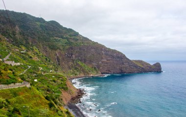 Koyu bulutlar, yüksek dağlar ve dik uçurumlarla kaplı bir sahil şeridi olan Madeiras 'ın kuzey kıyısındaki dramativ sahnesi. Öndeki yeşil tarlalarda ve Sao Jorge 'nin küçük köyünde. Büyük dalgalı mavi deniz.