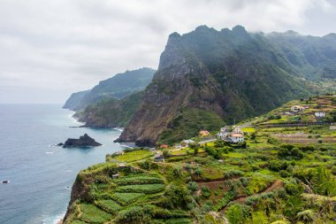 Koyu bulutlar, yüksek dağlar ve dik uçurumlarla kaplı bir sahil şeridi olan Madeiras 'ın kuzey kıyısındaki dramativ sahnesi. Öndeki yeşil tarlalarda ve Sao Jorge 'nin küçük köyünde. Büyük dalgalı mavi deniz.
