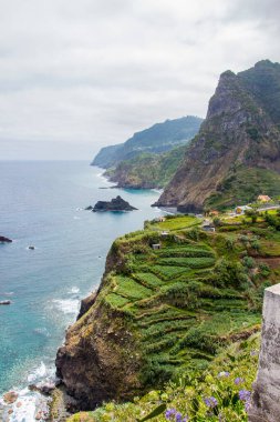 Koyu bulutlar, yüksek dağlar ve dik uçurumlarla kaplı bir sahil şeridi olan Madeiras 'ın kuzey kıyısındaki dramativ sahnesi. Öndeki yeşil tarlalarda ve Sao Jorge 'nin küçük köyünde. Büyük dalgalı mavi deniz.