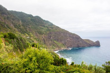 Koyu bulutlar, yüksek dağlar ve dik uçurumlarla kaplı bir sahil şeridi olan Madeiras 'ın kuzey kıyısındaki dramativ sahnesi. Öndeki yeşil tarlalarda ve Sao Jorge 'nin küçük köyünde. Büyük dalgalı mavi deniz.