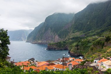 Koyu bulutlar, yüksek dağlar ve dik uçurumlarla kaplı bir sahil şeridi olan Madeiras 'ın kuzey kıyısındaki dramativ sahnesi. Öndeki yeşil tarlalarda ve Sao Jorge 'nin küçük köyünde. Büyük dalgalı mavi deniz.