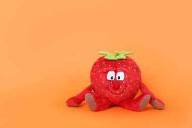 Parlak turuncu arka plandaki küçük çocuklar için peluş kırmızı çilek oyuncağı. Çocukluk konsepti. Boşluğu kopyala.