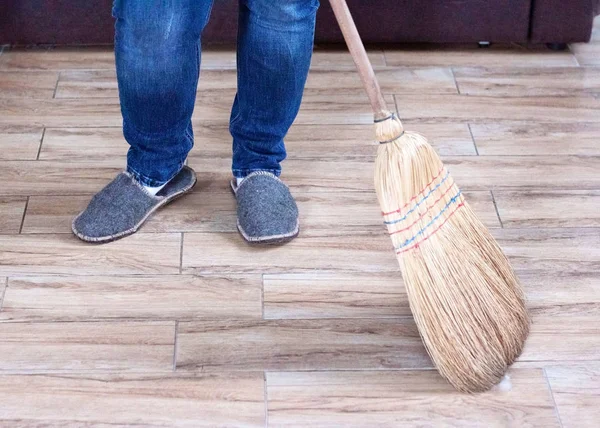 Kot pantolonlu ve terlikli bir adam süpürgenin yanında temiz bir zemini süpürür.