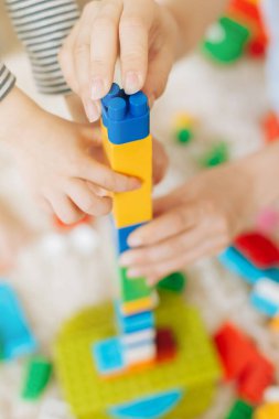 Anne ve kızı, yumuşak beyaz bir halıda renkli plastik bir inşaatçıyla birlikte oynuyorlar.