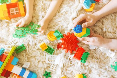 Anne ve kızı, yumuşak beyaz bir halıda renkli plastik bir inşaatçıyla birlikte oynuyorlar.