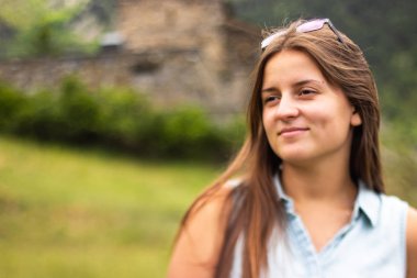 Uzun kahverengi saçlı genç bir kadın, yeşil tarlada, arkasında taş bir kilise var. Sığ alan derinliği