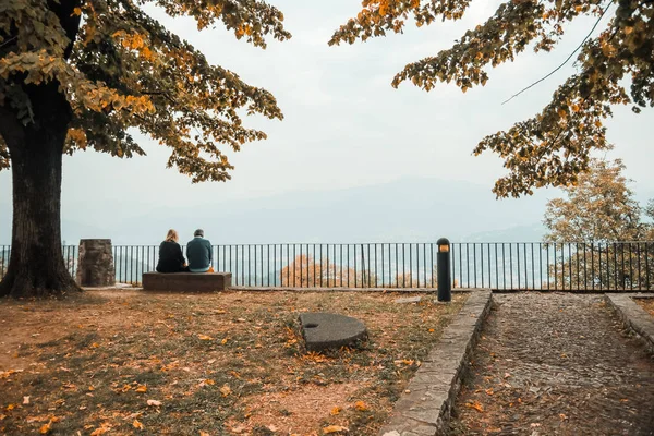 Dospělý pár sedí na lavičce v parku kolem stromů a před krásným výhledem na hory. — Stock fotografie