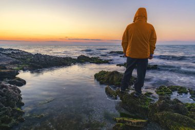 Kapüşonlu genç bir adam deniz kıyısında gün doğumuyla karşılaşır.