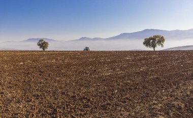 Traktör, toprağı dağların arka planına sürerek sürüyor.