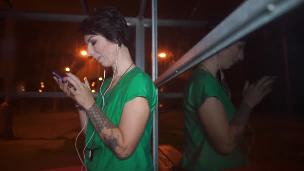 Девушка использует мобильный телефон, сидя на автобусной остановке в вечернее время — стоковое видео