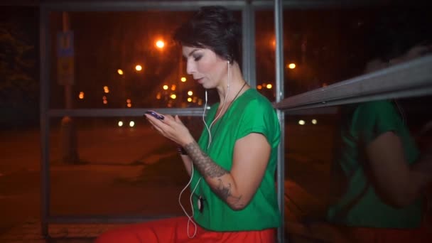 Lány használ egy okostelefon egy buszmegálló éjjel, a busz érkezik — Stock videók