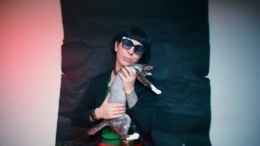 Siyah elbiseli bir kadın kollarındaki Devon Rex kedisini okşuyor..