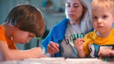 İki oğlu olan anne bir masaya oturur ve çizim yapar..