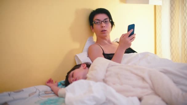 La mujer en la cama usa un teléfono móvil. Un hijo está durmiendo cerca. . — Vídeos de Stock