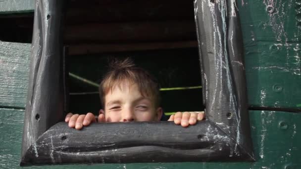 El niño se esconde en una casa de madera, mira por la ventana — Vídeo de stock