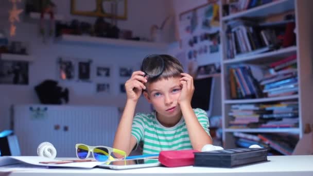 El niño se sienta en una mesa en la escuela, gira una lupa cerca de su cabeza — Vídeo de stock