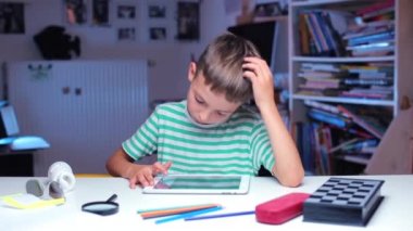 Bir çocuk sınıf masasında oturuyor, tablet ekranına parmağını sürtüyor.