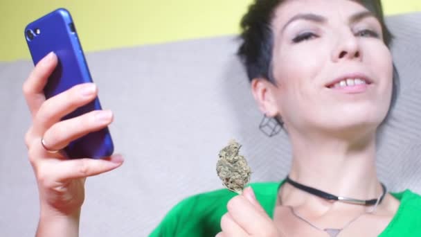 Ragazza sorride, dimostra un germoglio di cannabis nelle sue mani, tenendo un telefono — Video Stock