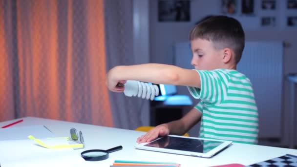 Niño utiliza la tableta mientras está sentado en una mesa, examina una bombilla de bajo consumo — Vídeos de Stock