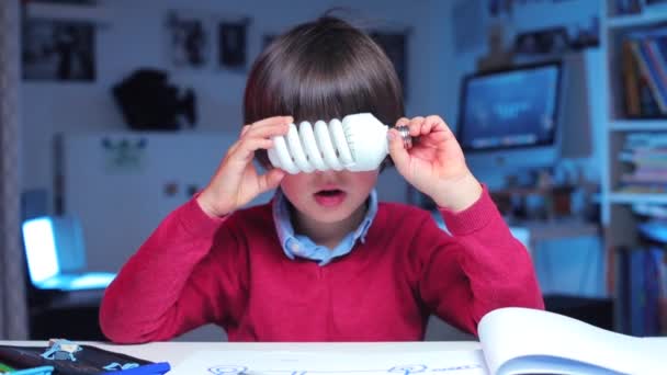 Niño juega con una bombilla de bajo consumo sentada en una mesa — Vídeos de Stock