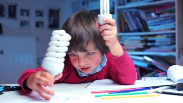 Niño con ropa roja sentado en una mesa examina las bombillas — Vídeos de Stock