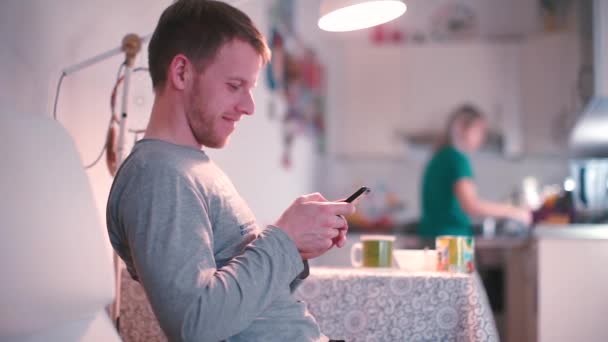 Un hombre se sienta en una mesa en la cocina, utiliza un teléfono inteligente . — Vídeos de Stock