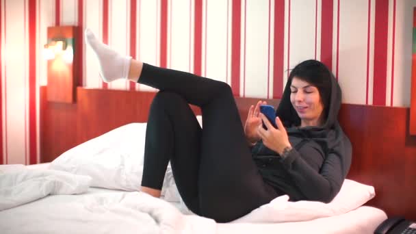 Menina em outerwear usa um smartphone em uma cama de hotel . — Vídeo de Stock