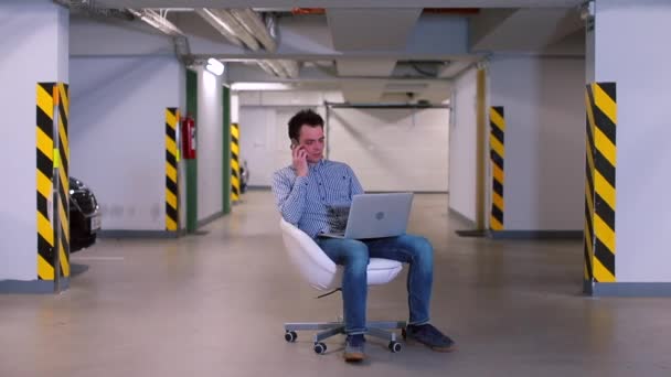 Un employé de bureau est assis sur une chaise dans un garage souterrain avec un ordinateur portable . — Video