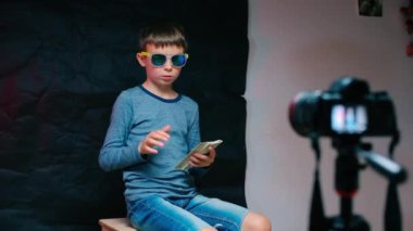 Gözlüklü bir çocuk video kameranın önünde bir sandalyeye oturur..