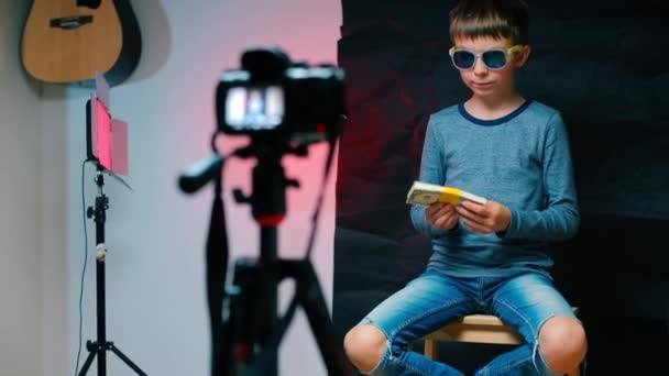 Мальчик сидит на стуле перед камерой и показывает деньги, заработанные на блоге . — стоковое видео