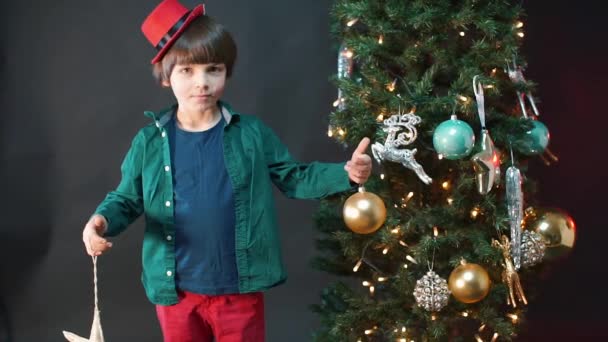 Un bambino con un cappello rosso si trova vicino a un albero di Natale . — Video Stock
