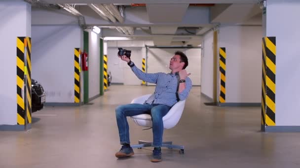 A man sits in an underground garage in an armchair, holds a camera in his hands. — ストック動画