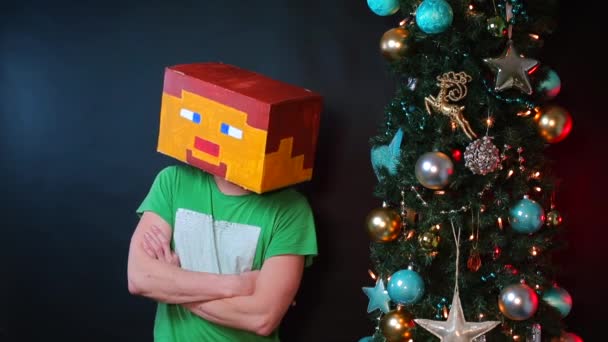 Man met een beschilderd masker met gevouwen armen op zijn borst zit bij de kerstboom. — Stockvideo