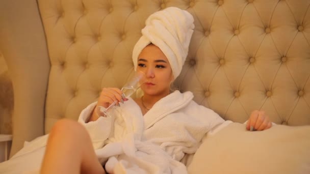 A noiva em um roupão de banho branco com uma toalha na cabeça fica na cama do hotel . — Vídeo de Stock
