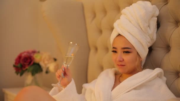 A young Asian woman drinks champagne while lying in bed. — Stockvideo