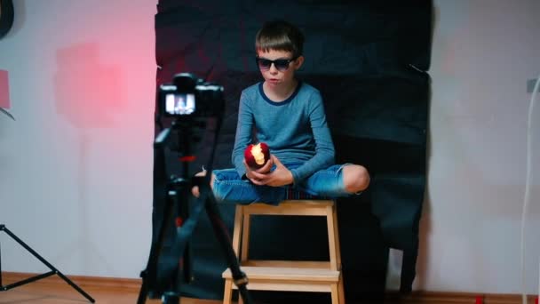 Un blogger con gli occhiali da sole si siede davanti a una videocamera e mangia una mela . — Video Stock