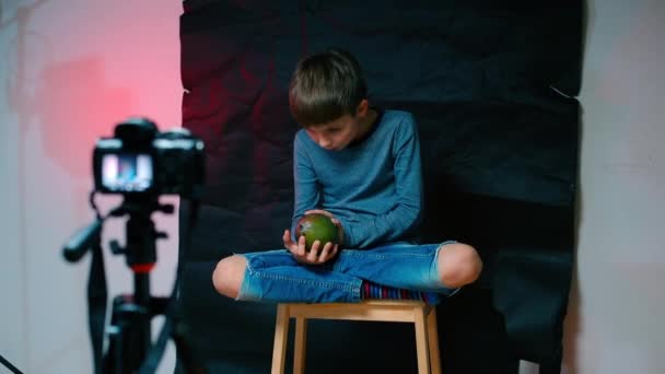 A boy sits in front of a video camera, holds a mango — Stockvideo