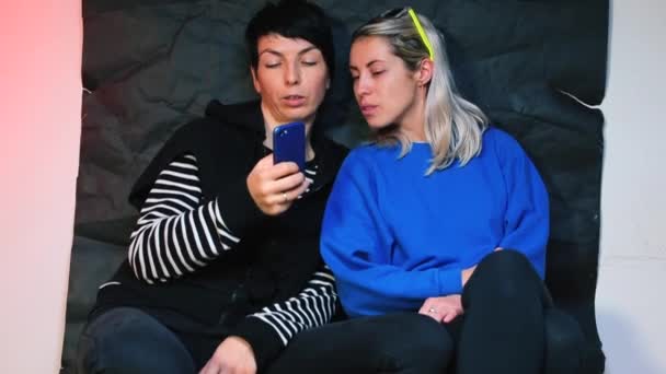 Dos hermanas están sentadas en un club nocturno, mirando la pantalla de un smartphone . — Vídeos de Stock