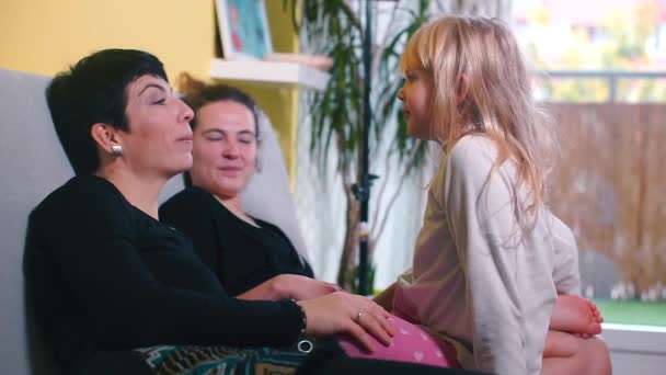 Duas mães LGBT com sua filha estão conversando no sofá em casa . — Vídeo de Stock