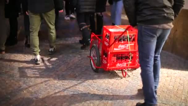 En man kör en vagn med röda lådor runt staden på kvällen bland människor — Stockvideo