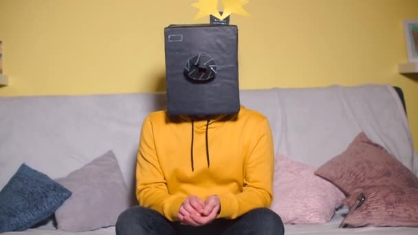 Un hombre con ropa amarilla enmascarada se sienta en un sofá con una máscara en la cabeza — Vídeos de Stock