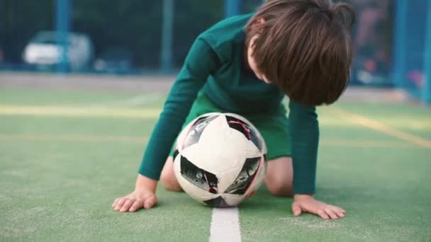 Αγόρι κάθεται πάνω από μια μπάλα ποδοσφαίρου στην παιδική χαρά — Αρχείο Βίντεο