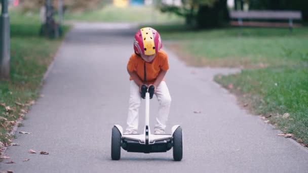 Un niño en una camiseta naranja monta un segway a lo largo del camino de un parque de la ciudad — Vídeos de Stock