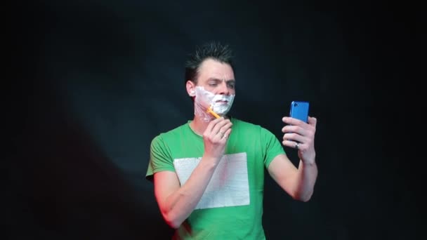 El video blogger se afeita mientras mira un teléfono inteligente en lugar de un espejo . — Vídeo de stock