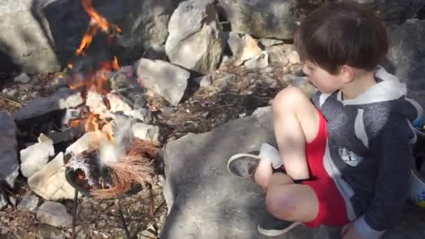 Niño dispara un fuego sentado en una piedra — Vídeos de Stock